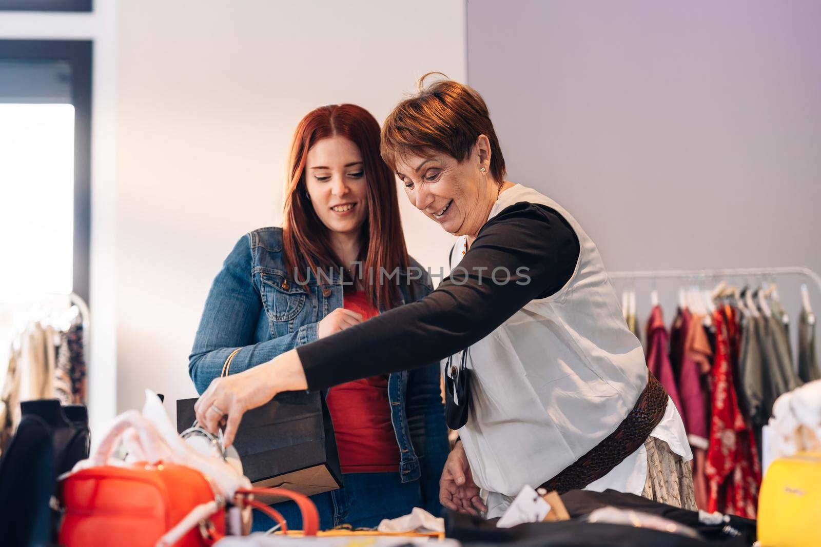 old woman and young woman buying clothes in a fashion shop. grandmother and granddaughter enjoying a shopping day. shopping concept. leisure concept. two smiling and happy people. Natural light, sunshine, clothes rack with colourful clothes, horizontal view, space for copying. casually dressed.