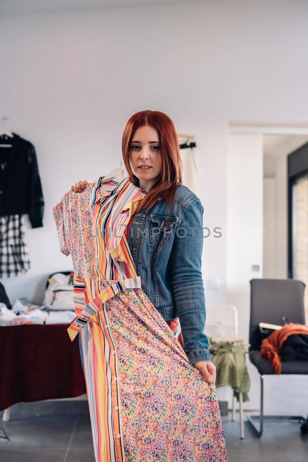 young woman trying on a dress in front of a mirror. red-haired girl shopping in a fashion shop. shopping concept. leisure concept. Natural light, sunbeams, display, clothes rack, clothes, vertical view, space to copy. customer in blue denim jacket, red t-shirt.