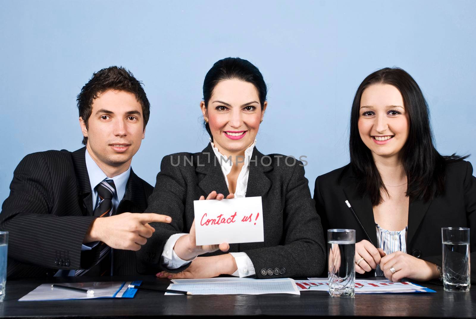 Group of three business people teamwork sitting in a line at table smiling and looking for you, a businesswoman holding a card with contact us handwritten displaying and her colleague man pointing to message