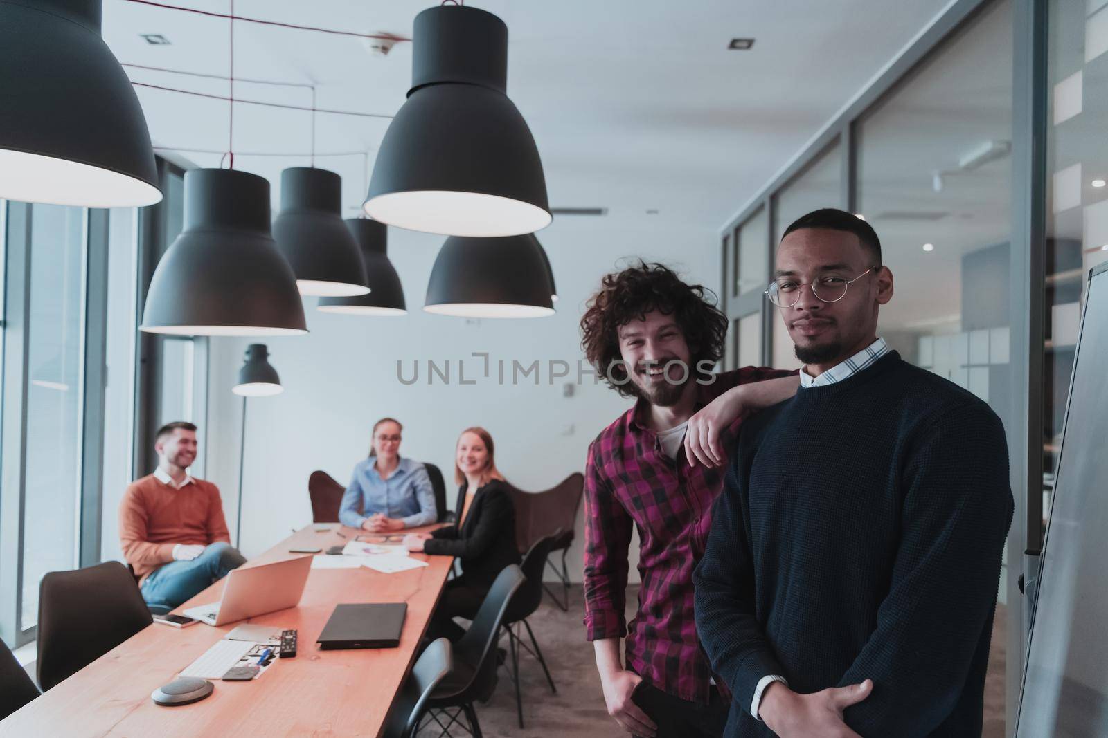 Portrait of two happy millennial male business owners in modern office. Two businessmen smiling and looking at the camera. Busy diverse team working in the background. Leadership concept. Head shot. High-quality photo