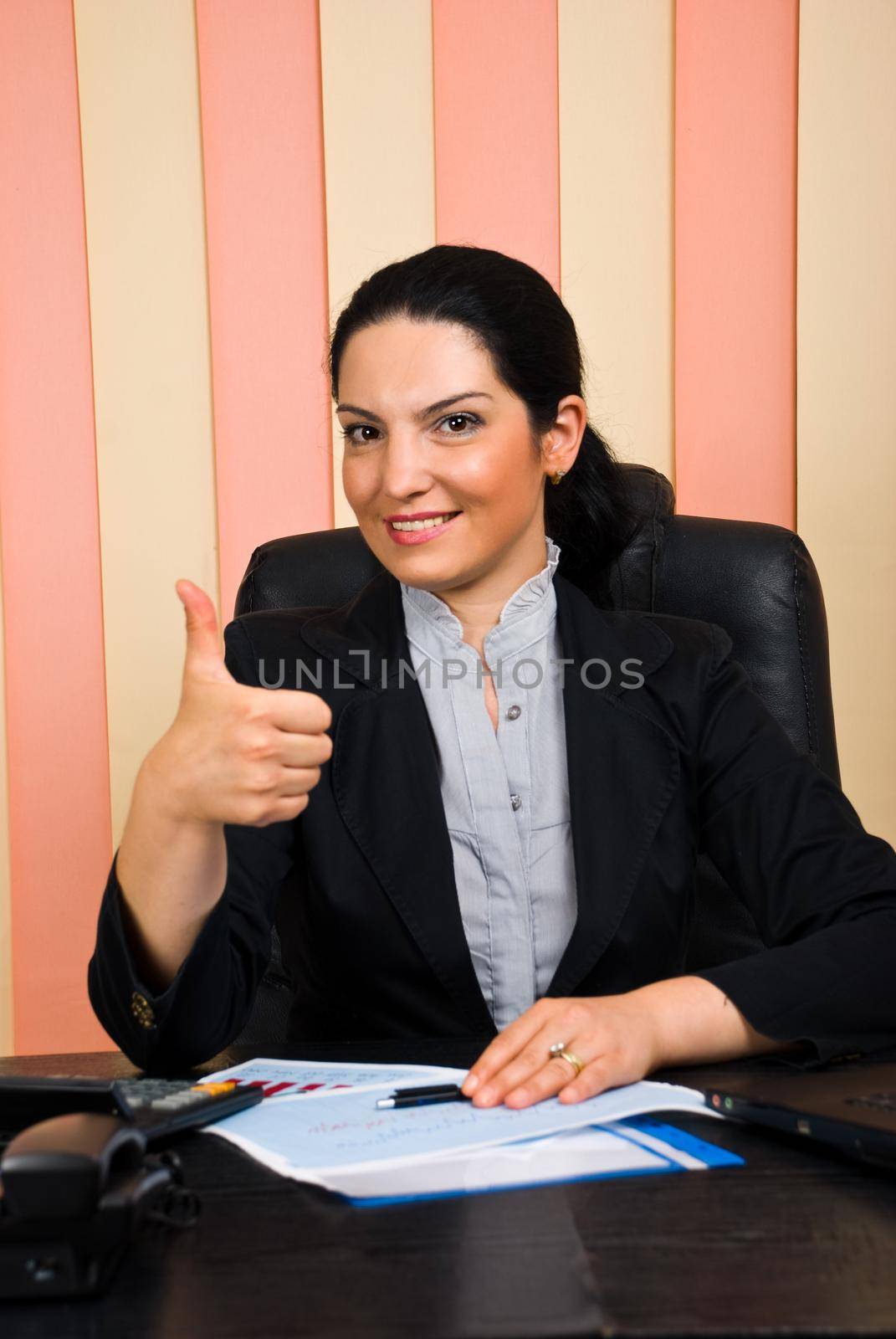 Elegant business woman sittiing on chair at desktop in her office and giving thumbs up