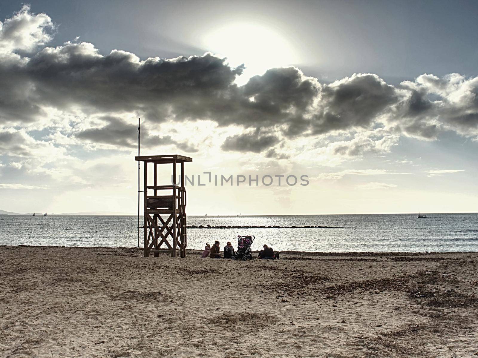 Family take a rest on sandy beach bellow lifeguard tower. Calm evening sea in bay. Palma de Mallorca, 26th of January 2020