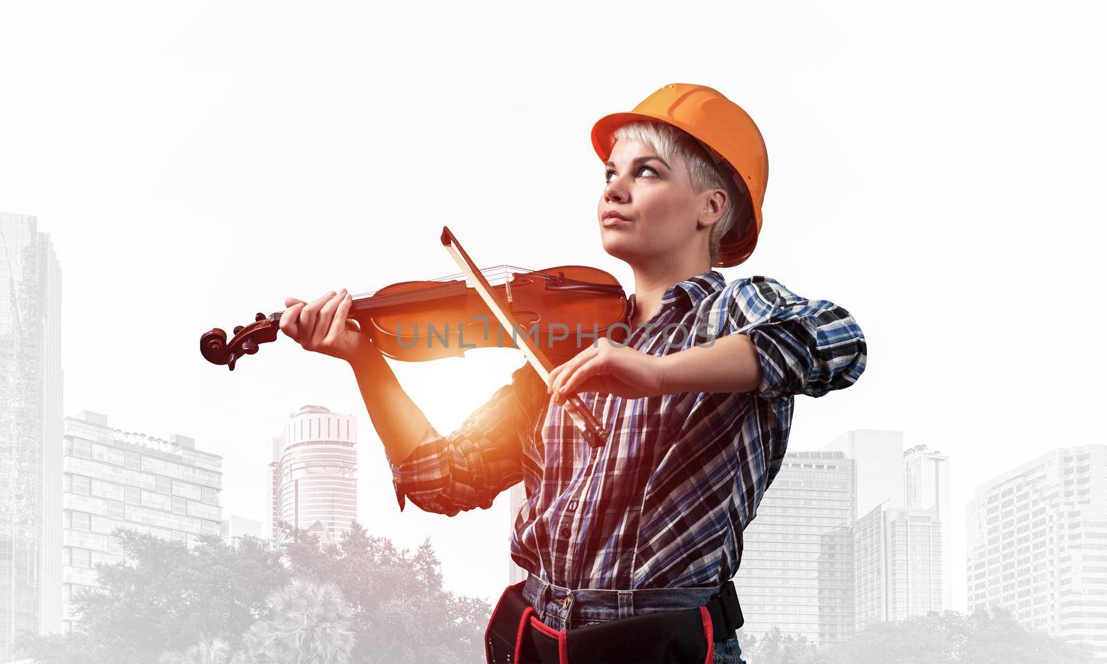 Beautiful young woman in safety helmet playing violin. Happy female builder in checkered blue shirt with instruments on foggy metropolis background. Industrial architecture and construction concept