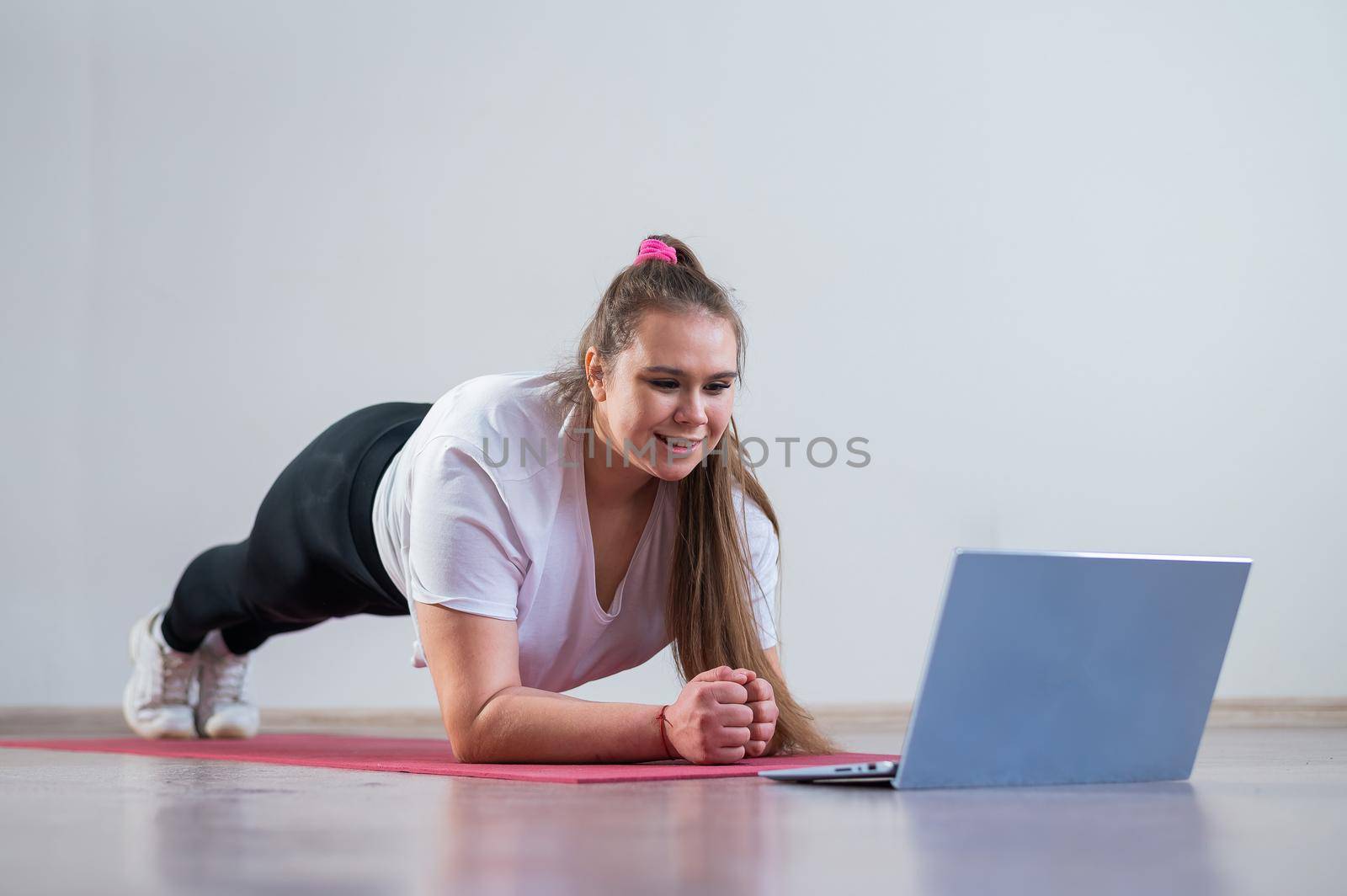 Young fat caucasian woman doing a plank on a sports mat. A cute plus size girl in sportswear is doing fitness exercises and watching an online tutorial on a laptop.