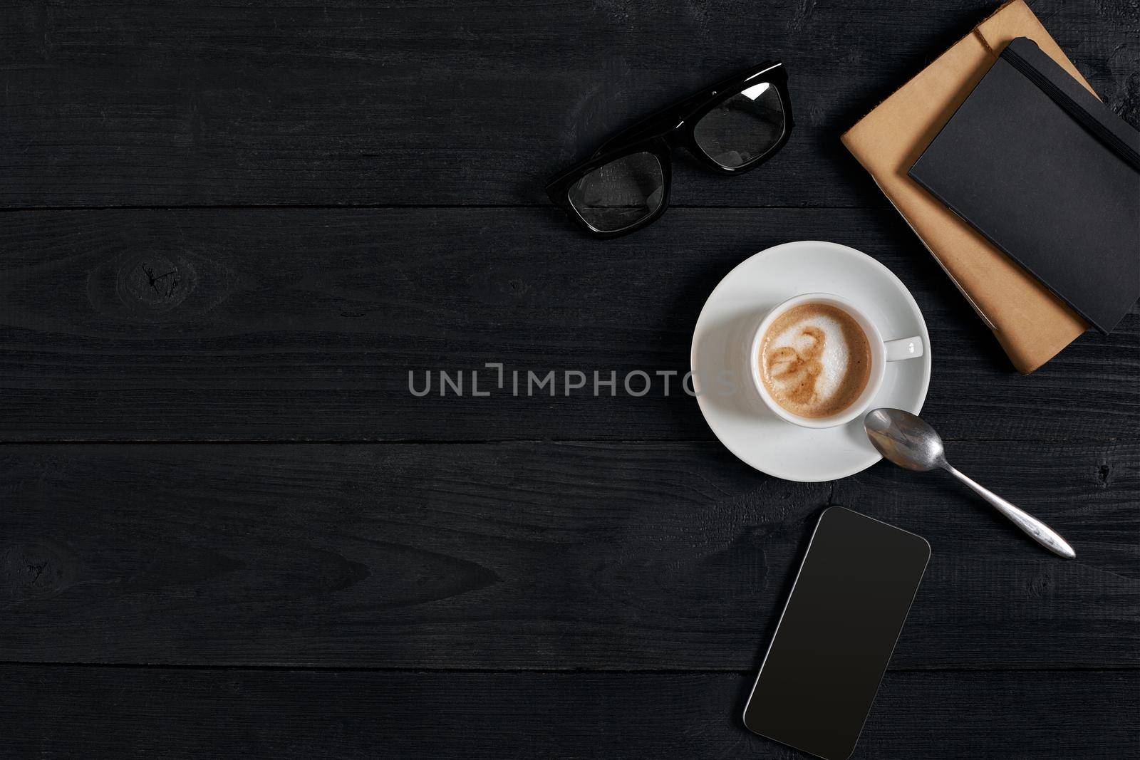 Above view of Smart phone with notebook and cup of latte coffee on black wooden background. Still life. Copy space. Flat lay. Top view