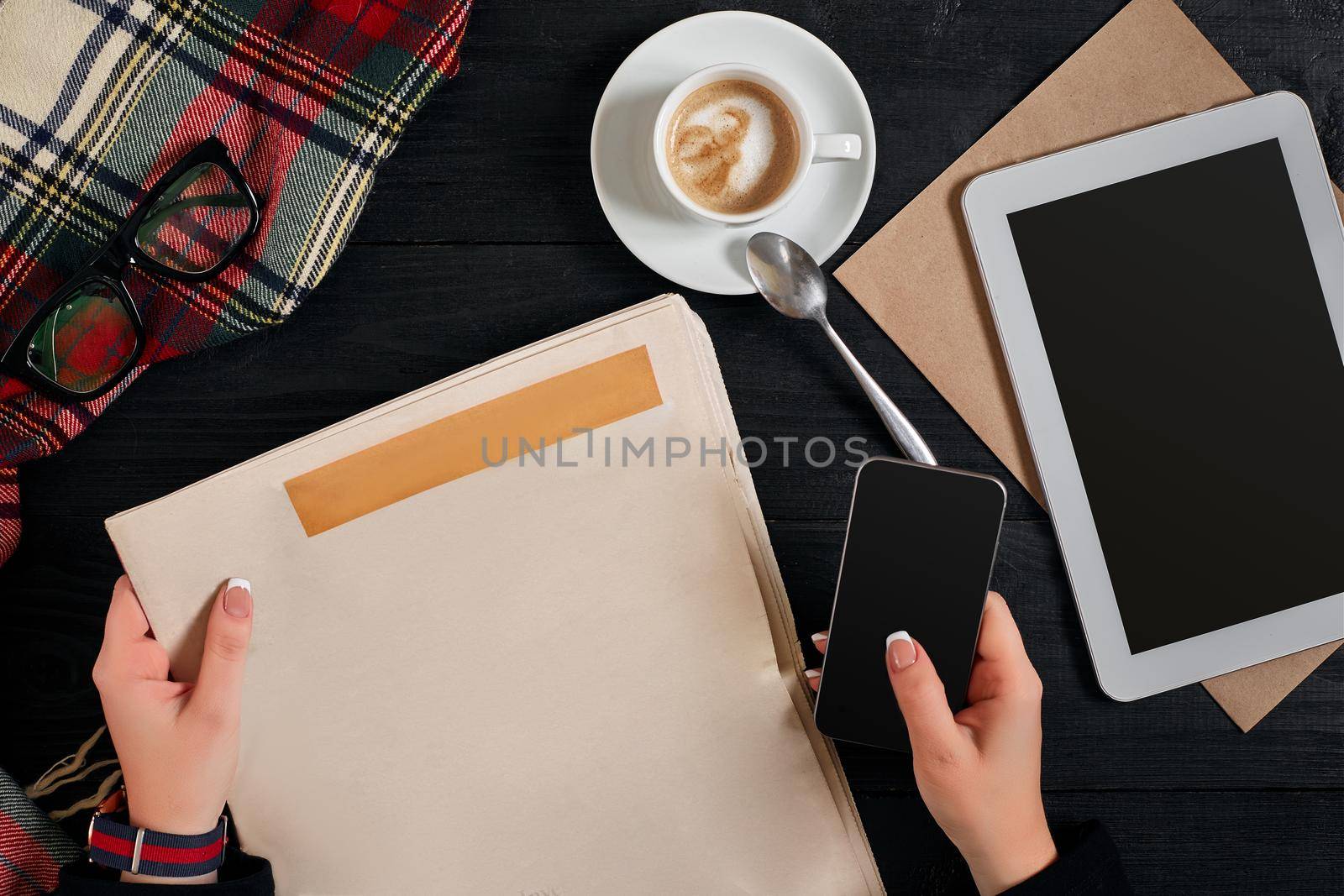 Newspaper and coffee cup, reading glasses, notebook, hands holding smartphone on a black wooden background. Stylish office desk. Autumn or Winter concept. Flat lay, top view. Still life. Copy space