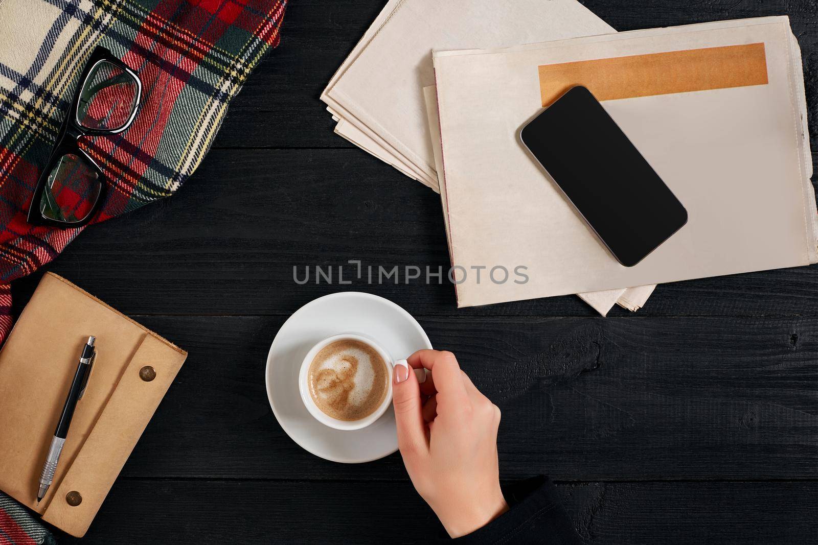 Newspaper and smartphone, reading glasses, notebook, hands holding coffee cup on a black wooden background. Stylish office desk. Autumn or Winter concept. Flat lay, top view. Still life. Copy space