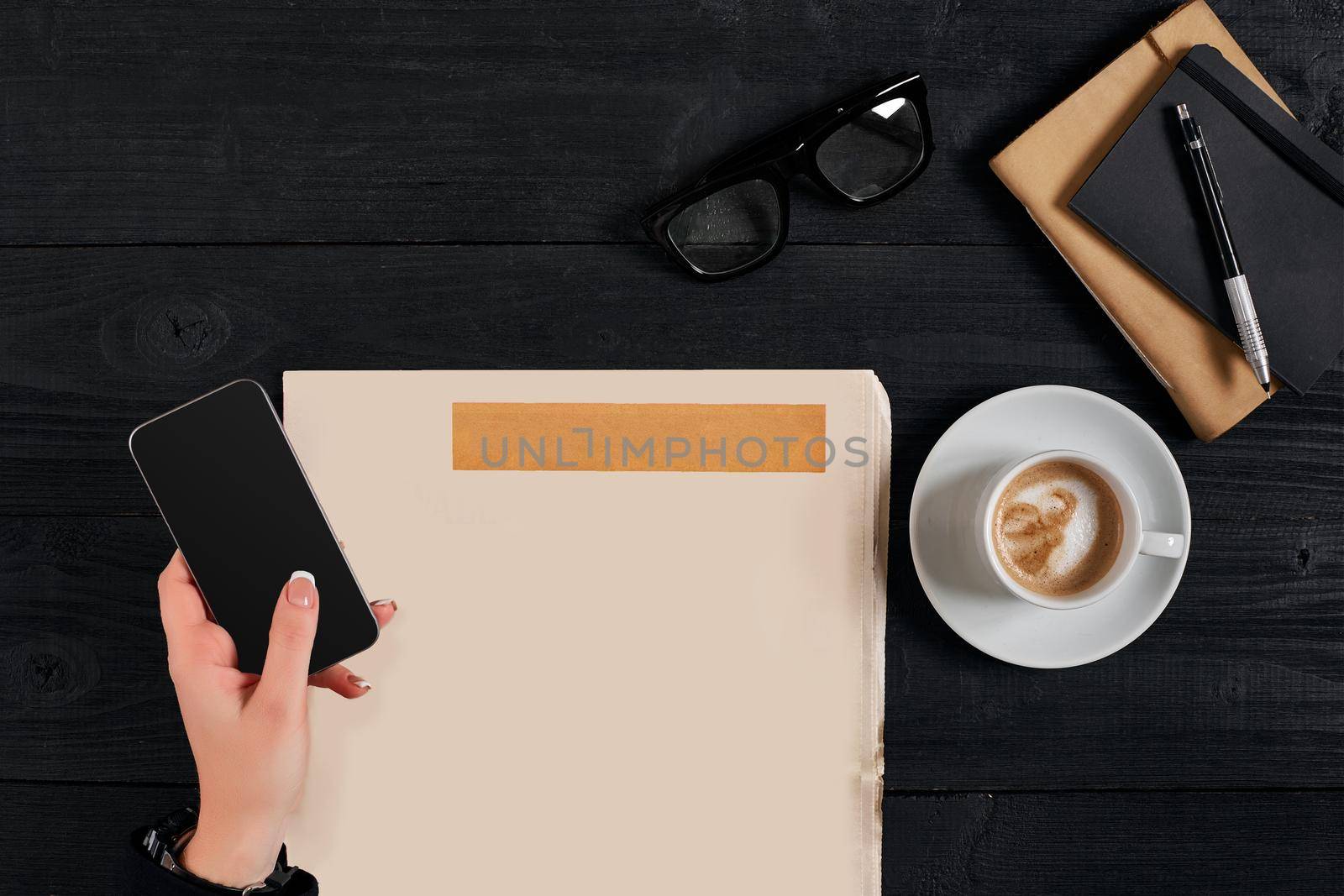 Newspaper and coffee cup, reading glasses, notebook, hands holding smartphone on a black wooden background. Stylish office desk. Autumn or Winter concept. Flat lay, top view. Still life. Copy space