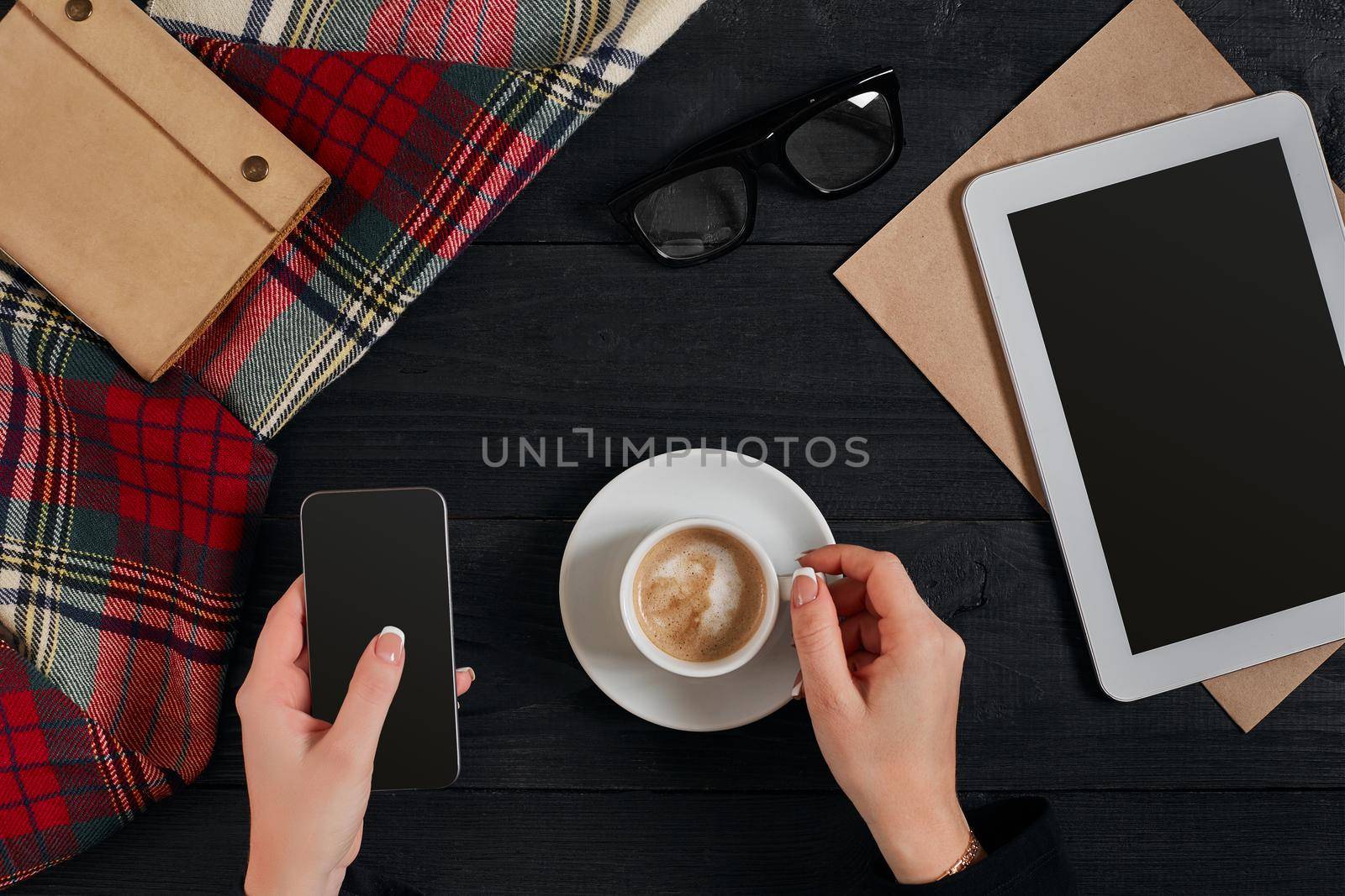 Women hands holding the phone with black screen above the table with a cup of coffee and tablet. Still life. Copy space. Flat lay. Top view