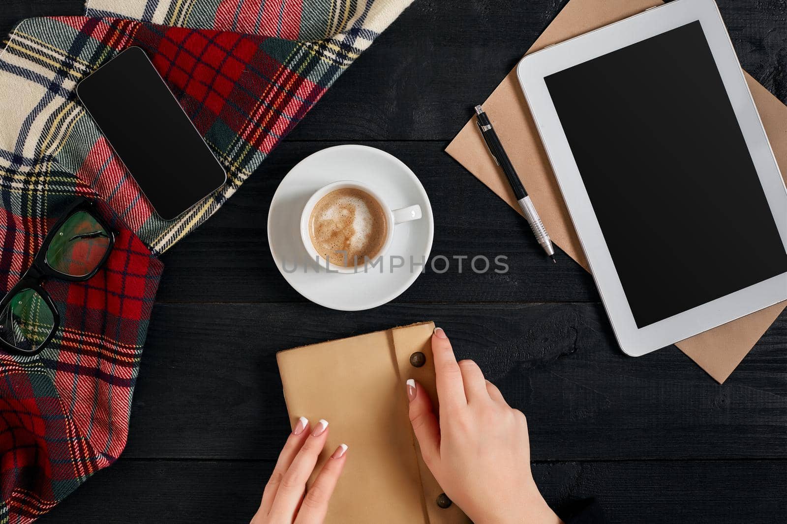 Young woman right hand writing on blank notebook on wood table with coffee cup, smartphone, and glasses beside in morning time. Top view. Still life. Flat lay