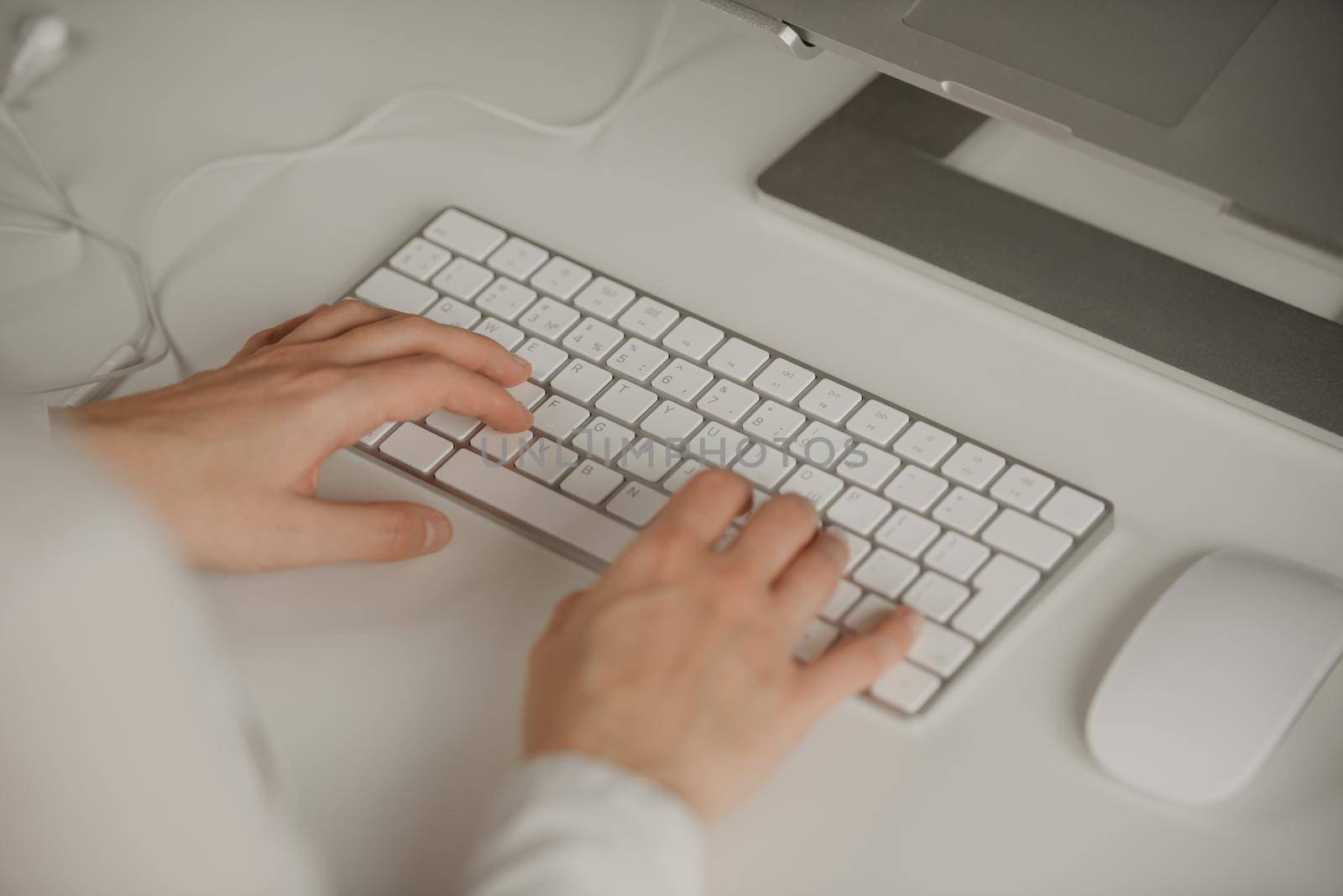 A workplace with an English keyboard. Close-up photo of woman’s hands typing on a wireless white keyboard. Aluminum laptop, headphones, keyboard, mouse.