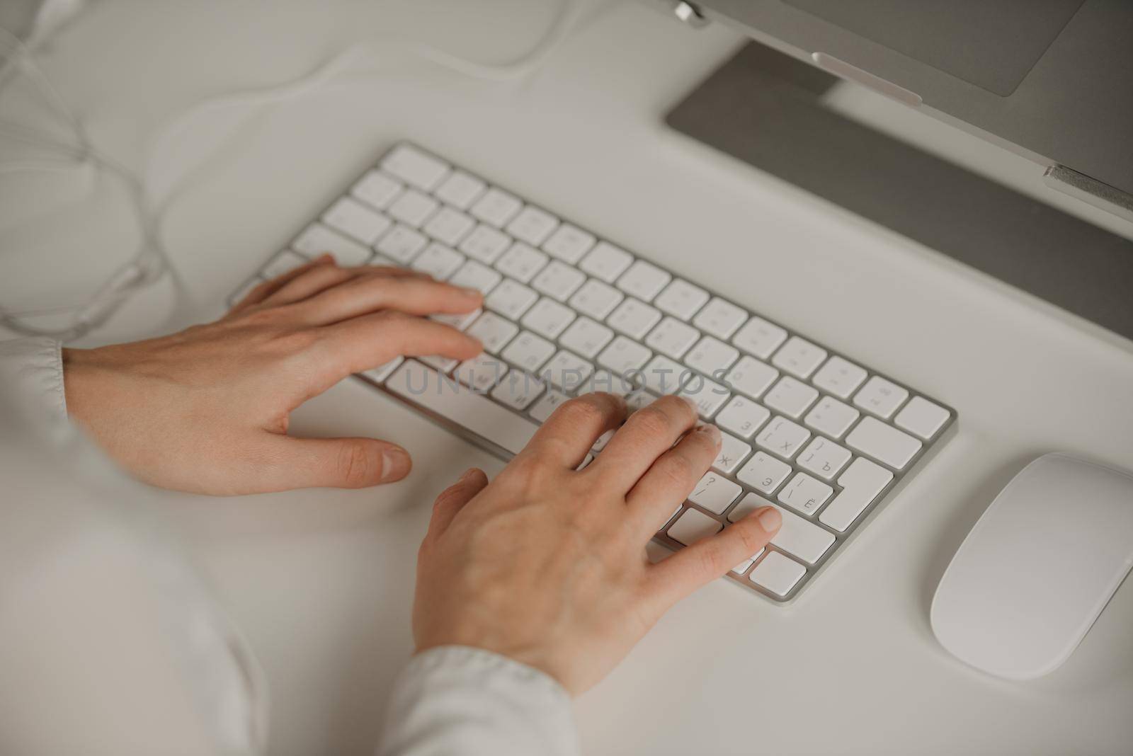 A workplace with an English-Russian keyboard. Close-up photo of woman’s hands typing on a wireless white keyboard. A girl working on a notebook. Aluminum laptop, headphones, keyboard, mouse