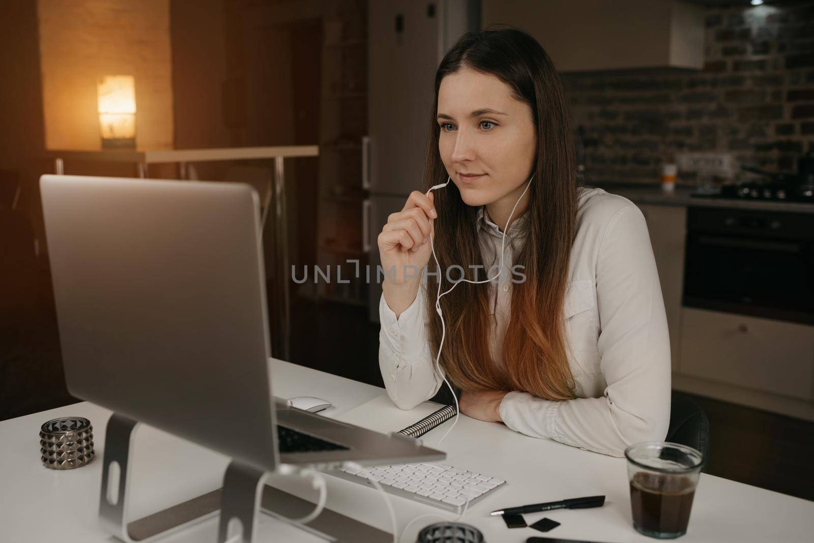 Remote work. A caucasian woman with headphones working remotely online on her laptop. A girl in a white shirt discussing business with her colleagues through a video call at her cozy home workplace.