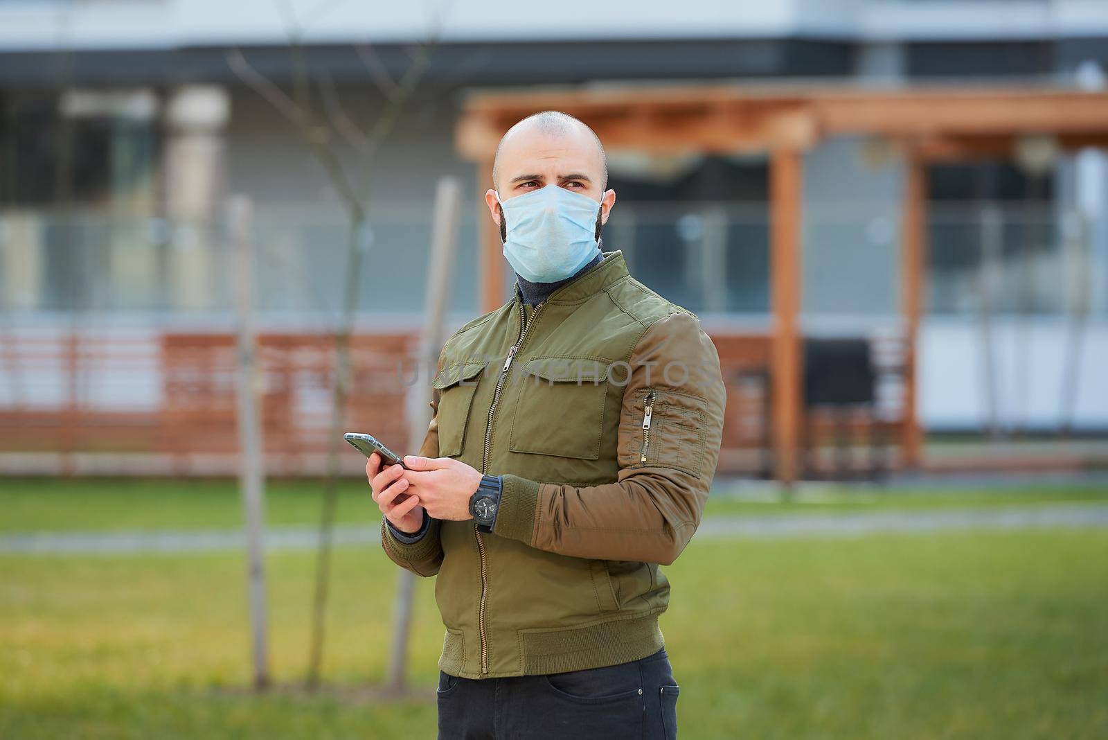 A bald man in a medical face mask to avoid the spread coronavirus checking his smartphone in the cozy street. A guy with a beard looking straight wears a face mask against COVID 19.