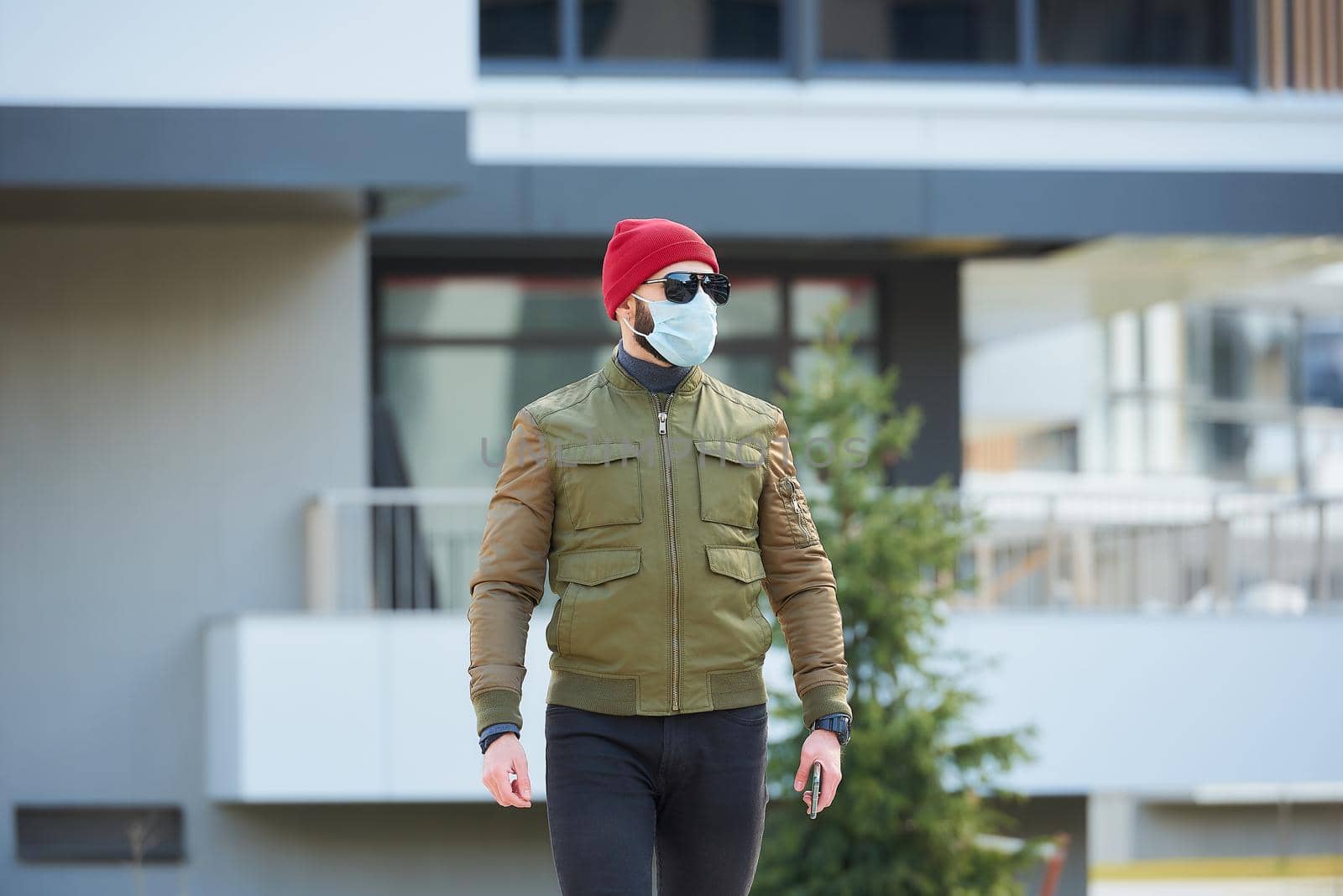 A man in a medical face mask to avoid the spread coronavirus holding his smartphone in a cozy street. A guy standing wears a red cap, sunglasses, and a face mask against COVID 19.