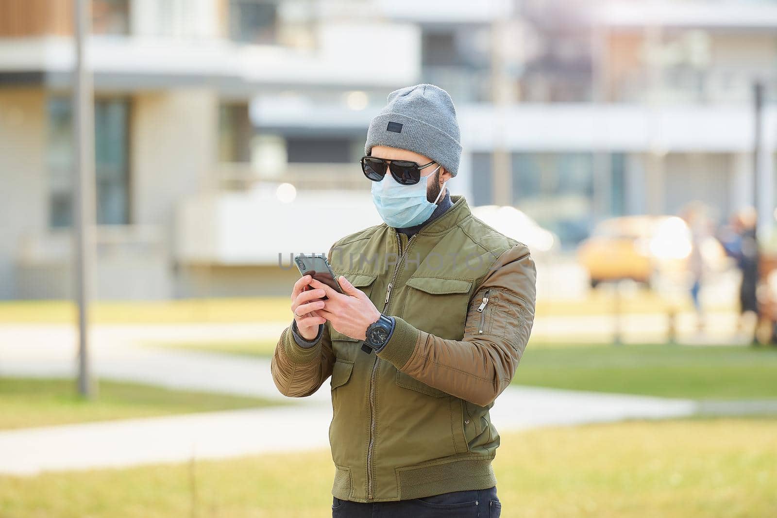 A man in a medical face mask to avoid the spread coronavirus holding his smartphone in the cozy street. A guy wears a gray cap, sunglasses, and a face mask against COVID 19.