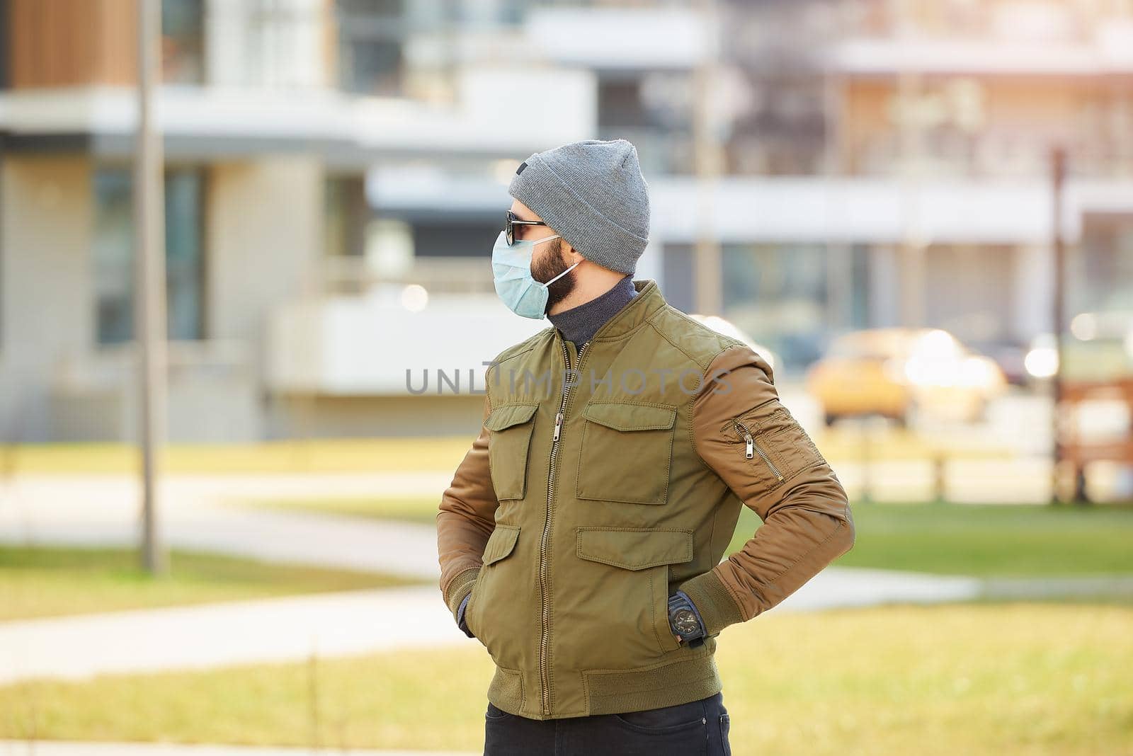 A man in a medical face mask to avoid the spread coronavirus waiting in the cozy street. A guy dropping his hands in his pockets wears a cap, sunglasses, and a face mask against COVID 19.