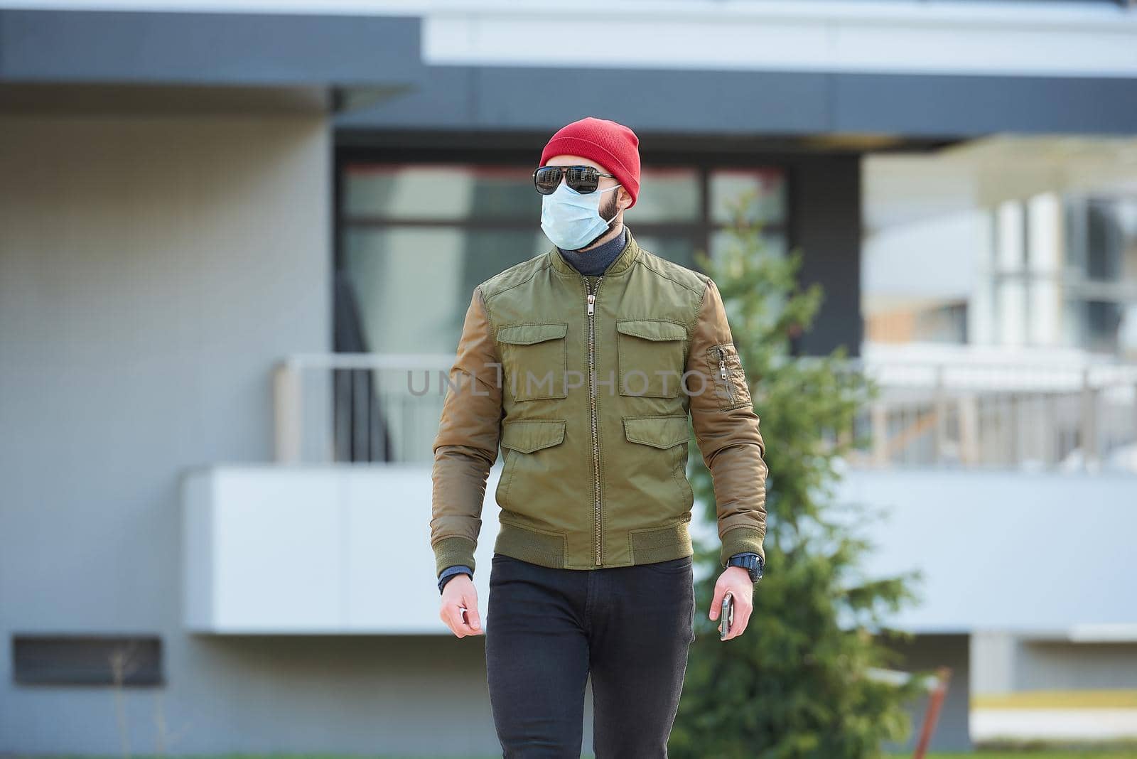 A man in a medical face mask to avoid the spread coronavirus holding his smartphone in a cozy street. A guy marching wears a red cap, sunglasses, and a face mask against COVID 19.