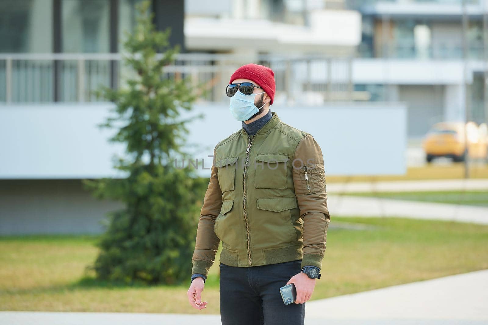 A man in a medical face mask to avoid the spread coronavirus holding his smartphone in a cozy street. A guy waiting wears a red cap, sunglasses, and a face mask against COVID 19.