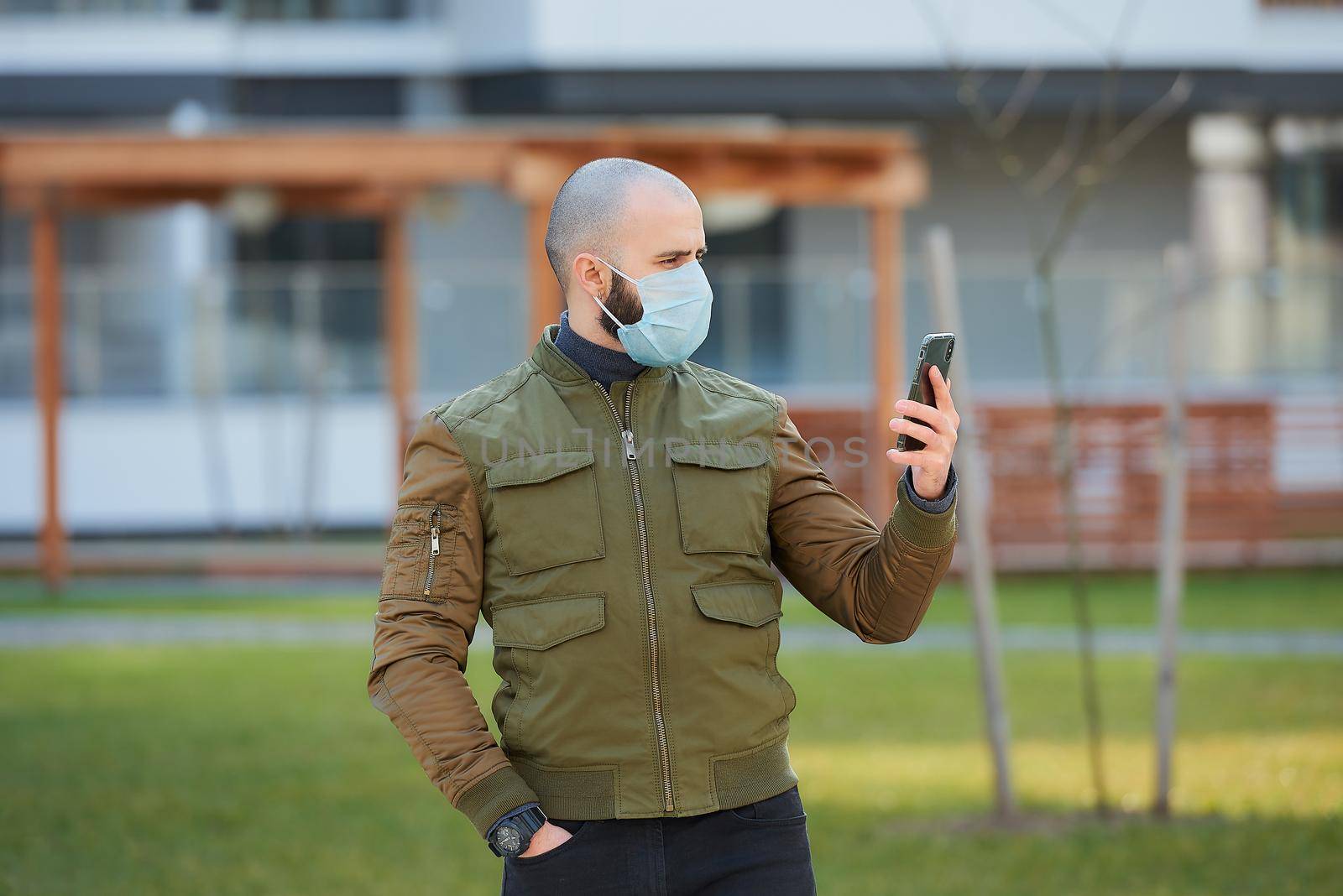 A bald man in a medical face mask to avoid the spread coronavirus checking his smartphone in the cozy street. A guy with a beard standing turned right wears a face mask against COVID 19.