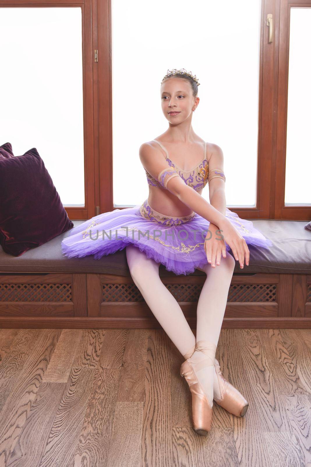 Full length portrait of a beautiful little ballerina in a performance purple dress sitting by the window and dreaming to become professional ballet dancer