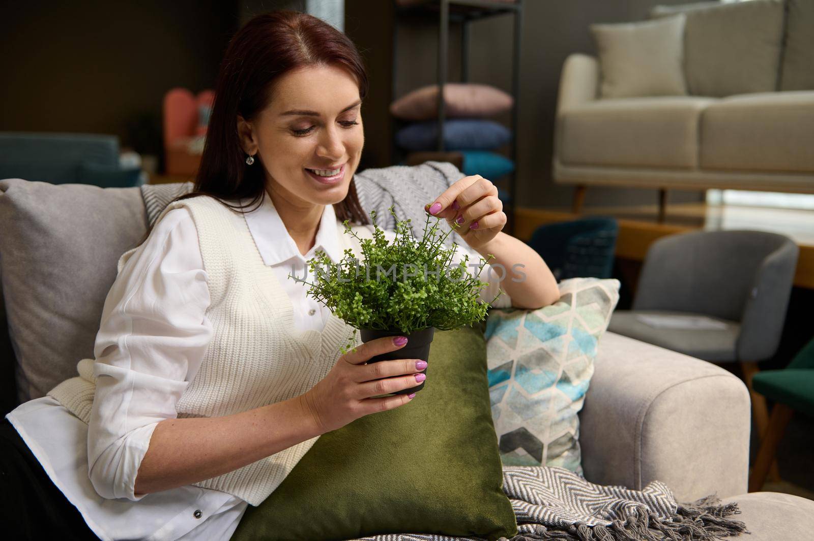 Beautiful middle-aged Caucasian woman holding ornamental plants sitting on a stylish modern comfortable sofa while shopping in the upholstered furniture store. Interior Design and home improvement