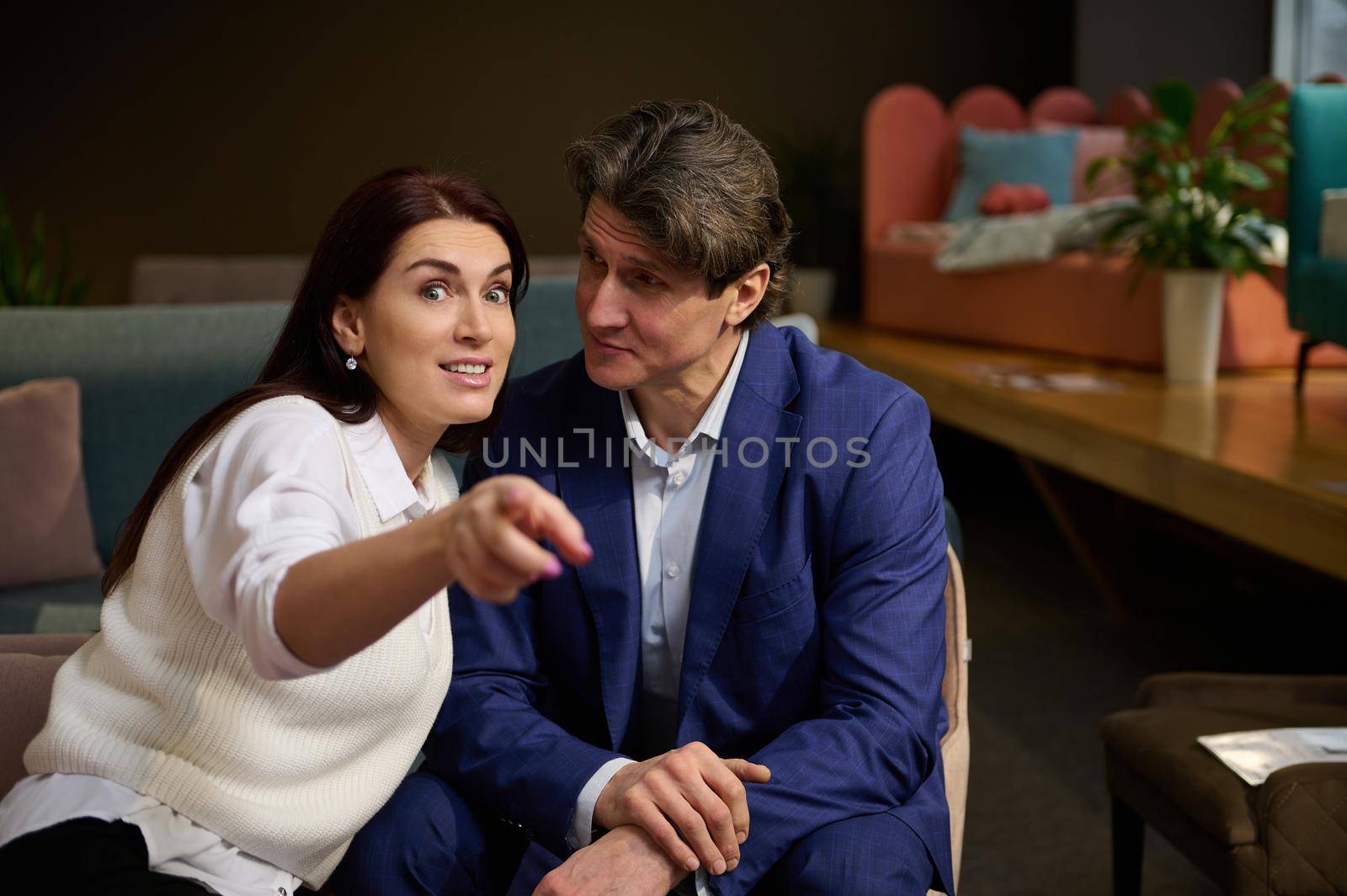 Handsome Caucasian man looking at his wife who is pointing on home goods in furniture shopping mall. Charming European woman with her husband looking for new sofa. Home improvement, interior design