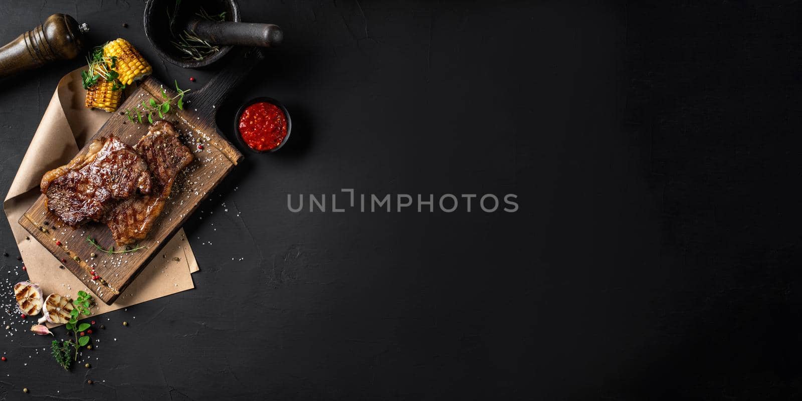 Fried steaks with herbs and spices on wooden board, parchment, garlic, corn, sprouts, pepper, salt, spice chopper, sauce. Black background. Top view. by nazarovsergey