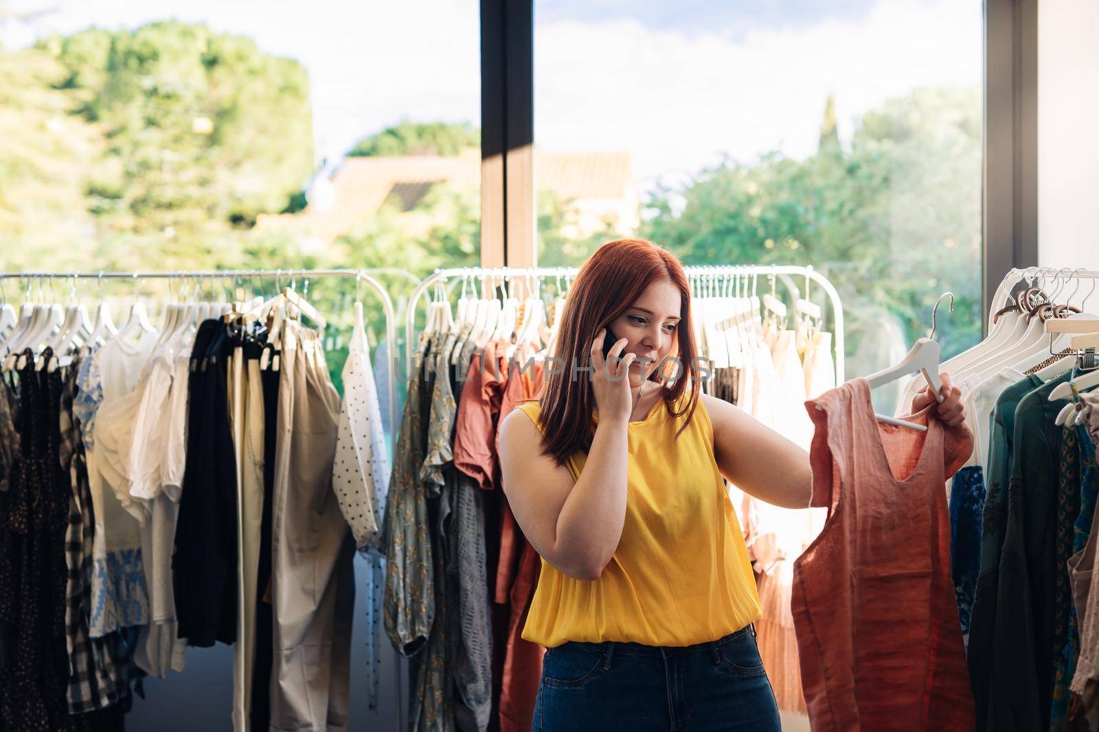 Young red-haired business woman buying clothes in a fashion shop. talking on mobile phone. shopping concept. Natural light, sun rays, display with clothes, clothes rack, clothes, vertical view. dressed in yellow t-shirt.