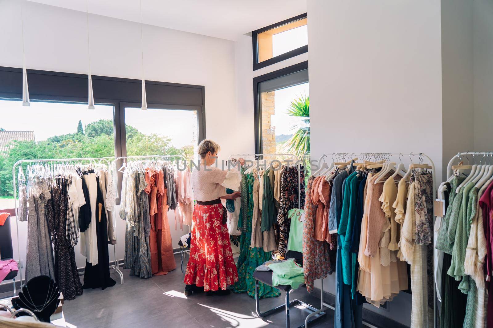 mature blonde woman, happy and enjoying a day of shopping, dressed in a blue denim jacket and a red t-shirt, choosing a pink dress from a rack, shopping in a fashion shop clothes. shopping concept. natural light from the shop window, sun rays, display with clothes, rack, shop window, clothes, Horizontal.