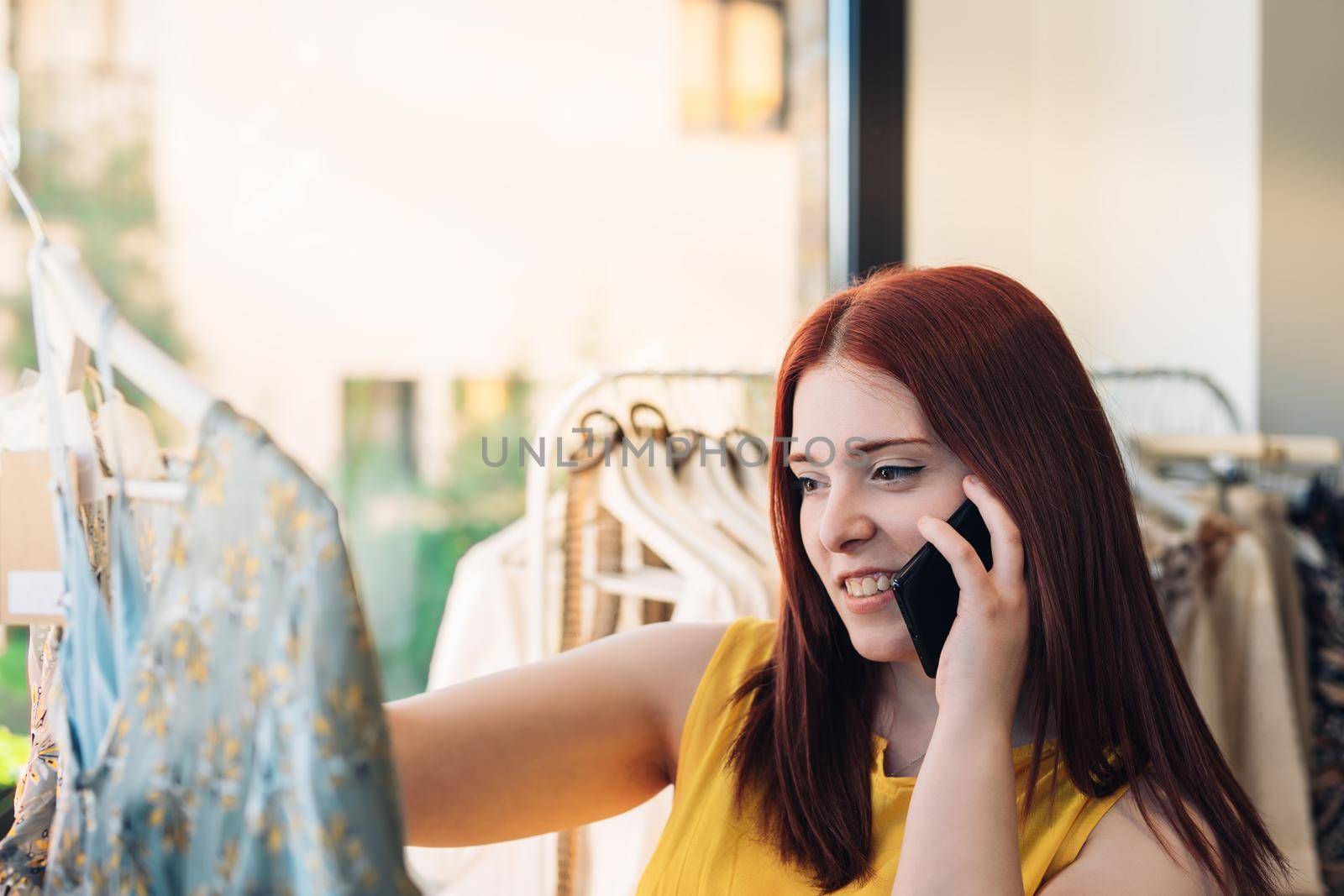 young red-haired woman shopping for clothes in a fashion shop. female talking on mobile phone. shopping concept. by CatPhotography