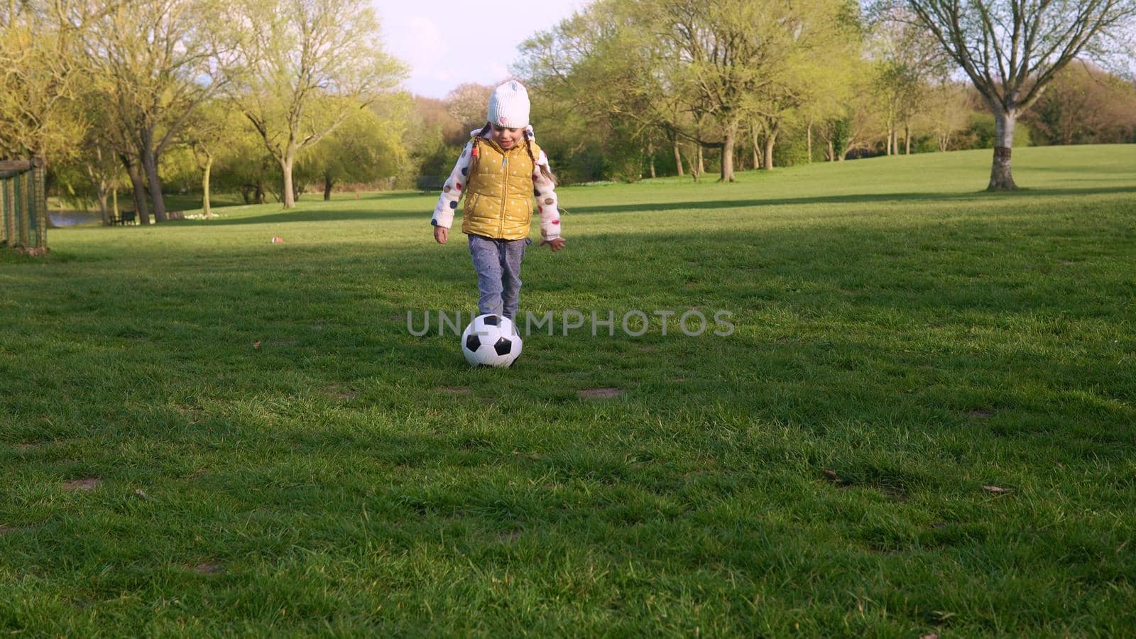 Happy Family Of Children Having Fun In Spring Park. Little Kid Run. Child Girl Dribbles Black White Classic Soccer Ball On Green Grass. People Playing Football. Childhood, Sport, Championship Concept by mytrykau