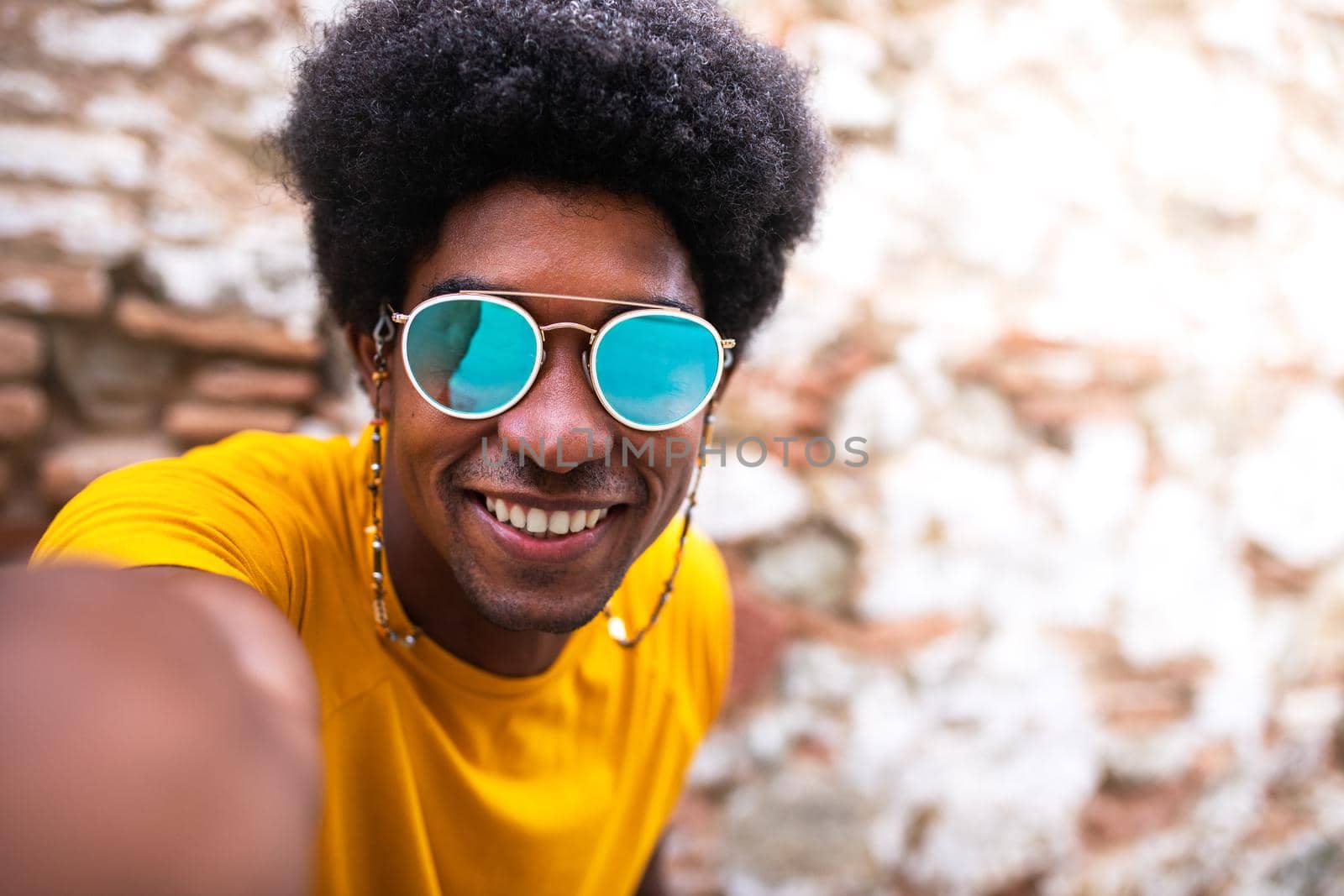 Happy, smiling young African American man taking selfie looking at camera. Copy space. Brick wall background. by Hoverstock