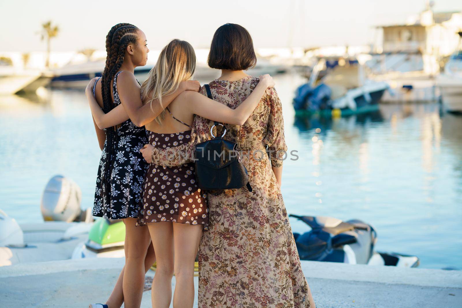 Back view of unrecognizable multiracial female friends in dresses hugging while standing on quay near river with vessels on summer day