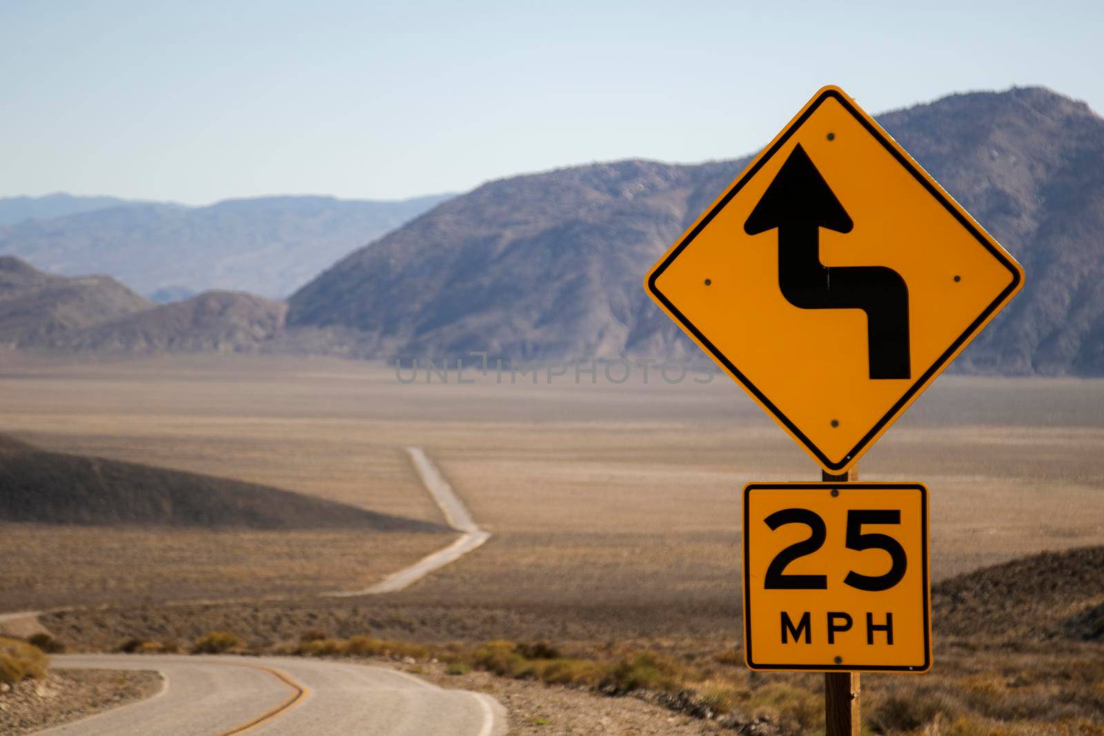 Long road in a desert with a curves sign in the foreground and some mountains in the background