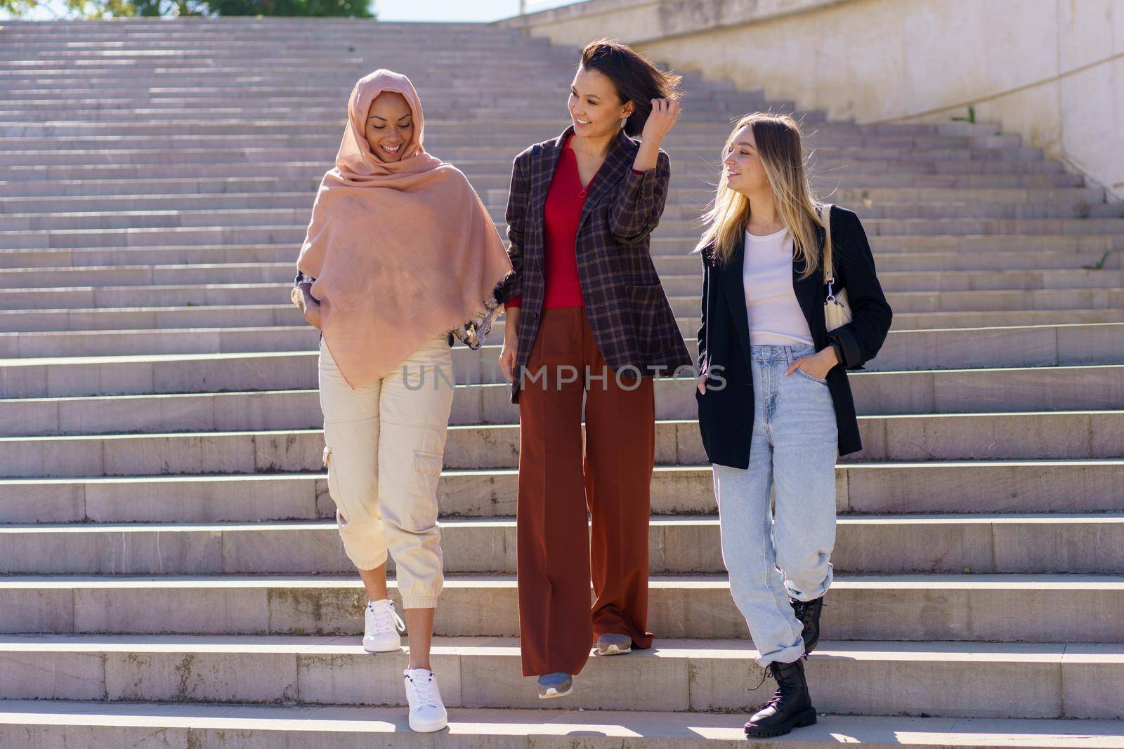 Confident young happy multiracial women students in casual outfits and hijab smiling while walking downstairs in university campus on sunny day