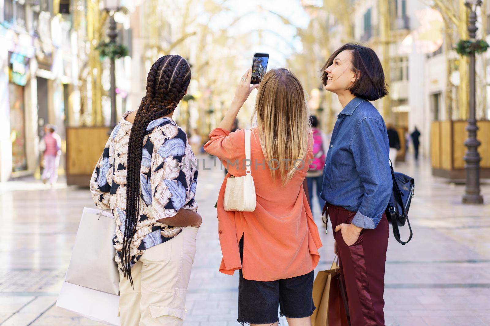 Group of diverse female friends standing together on blurred city street and taking selfie on mobile phone after shopping