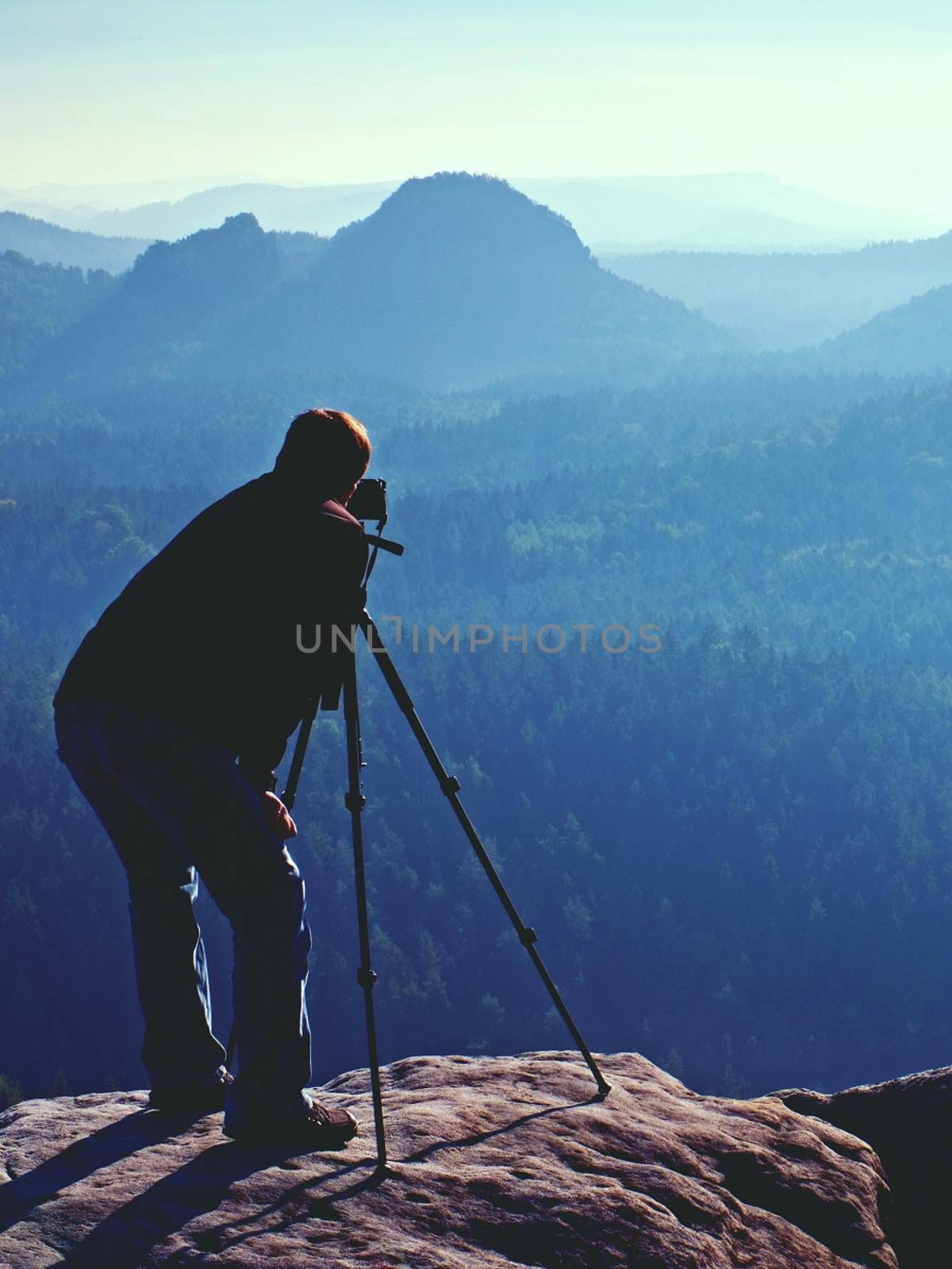 Hiker and photo enthusiast stay with tripod on cliff and thinking. Dreamy fogy landscape, blue misty sunrise in a beautiful valley below