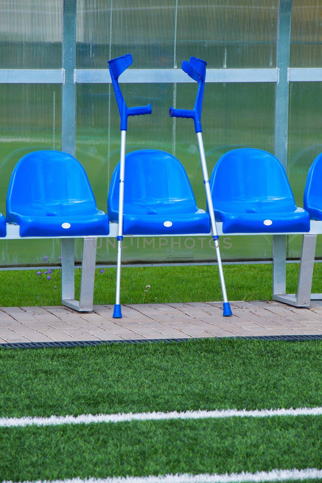 Crutch at blue plastic seats on outdoor stadium players bench, chairs with new paint below transparent plastic roof. 