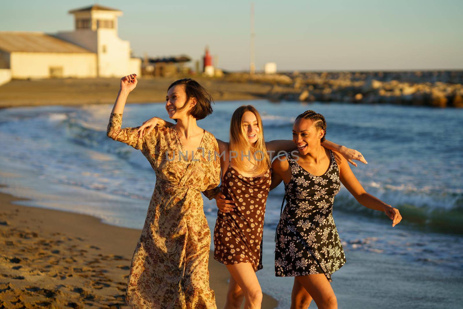Delighted multiracial women embracing each other while strolling on sandy seashore and enjoying summer sunset together