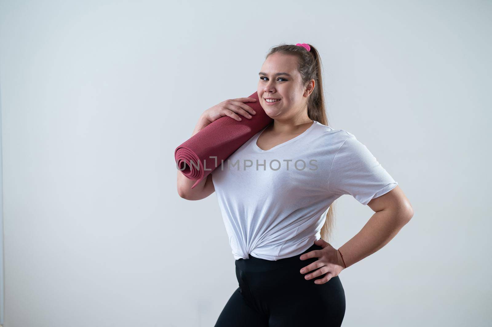 Young fat caucasian woman holding a sport mat. Charming plus size model in sportswear stands on a white background.