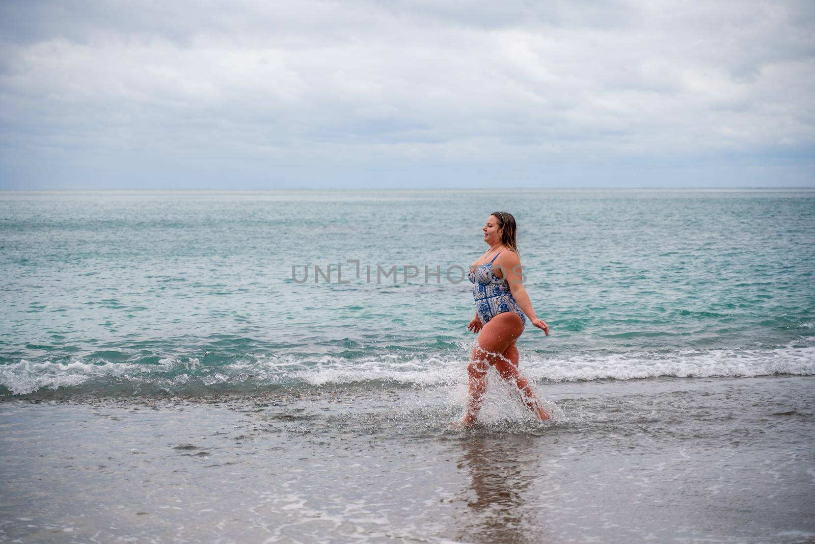 A plump woman in a bathing suit enters the water during the surf. Alone on the beach, Gray sky in the clouds, swimming in winter. by Matiunina