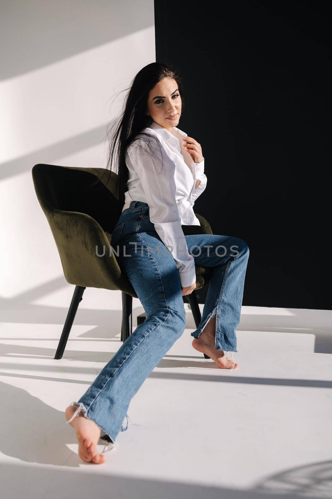 Pretty woman has photsession in studio. Sexy girl in denim and white shirt. Barefoot woman. Light from the sun makes a shadow on a white background.