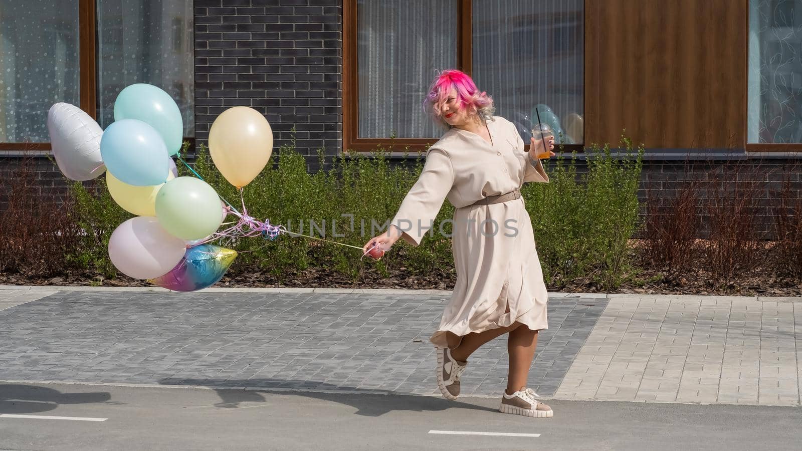 Woman in colored hair walks with an armful of balloons and drinks a refreshing beverage.
