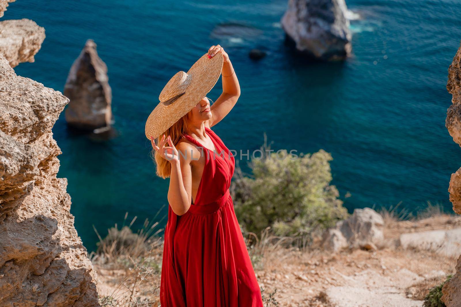 A woman in a flying red dress fluttering in the wind and a straw hat against the backdrop of the sea