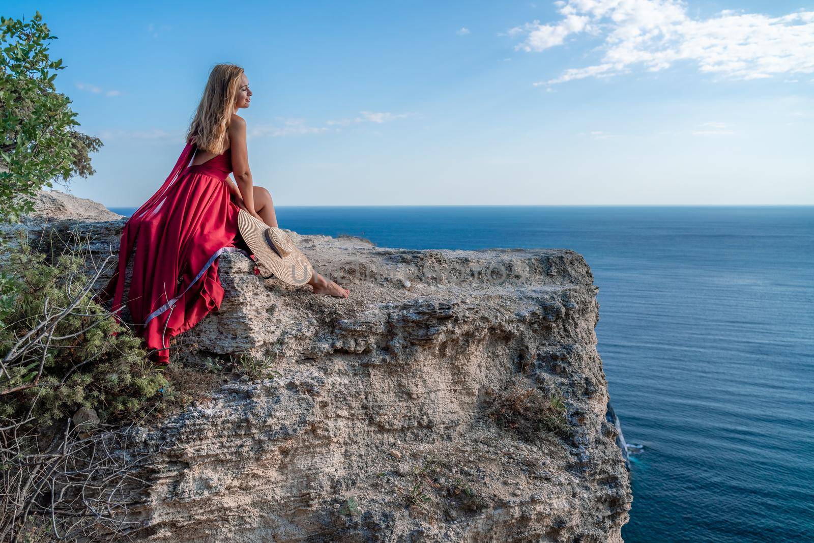 A girl with flowing hair in a long red dress sits on a rock above the sea. The stone can be seen in the sea. by Matiunina