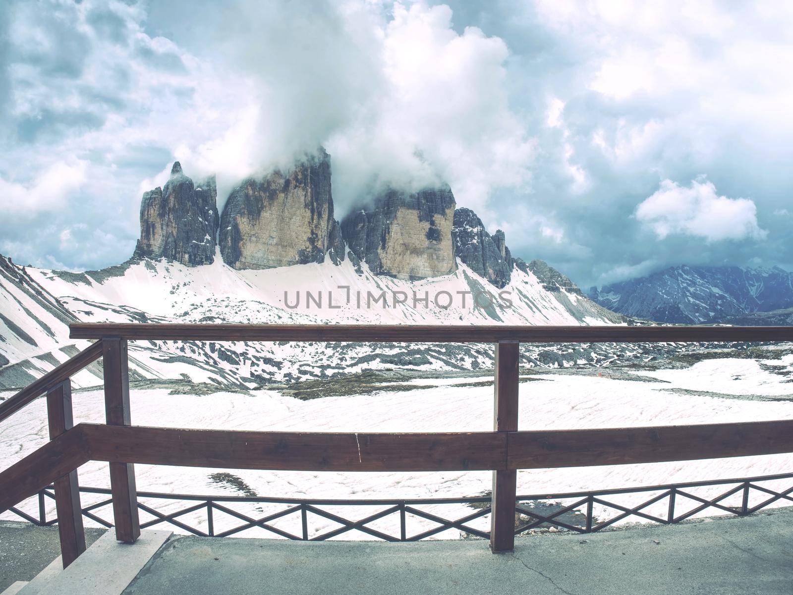 Breathtaking view from mountain hut terrace to symbol of Italian Dolomites - Tre Cime. High rocky ridge touching  clouds above snowy base. Tre Cime di Lavaredo National Park, Italy