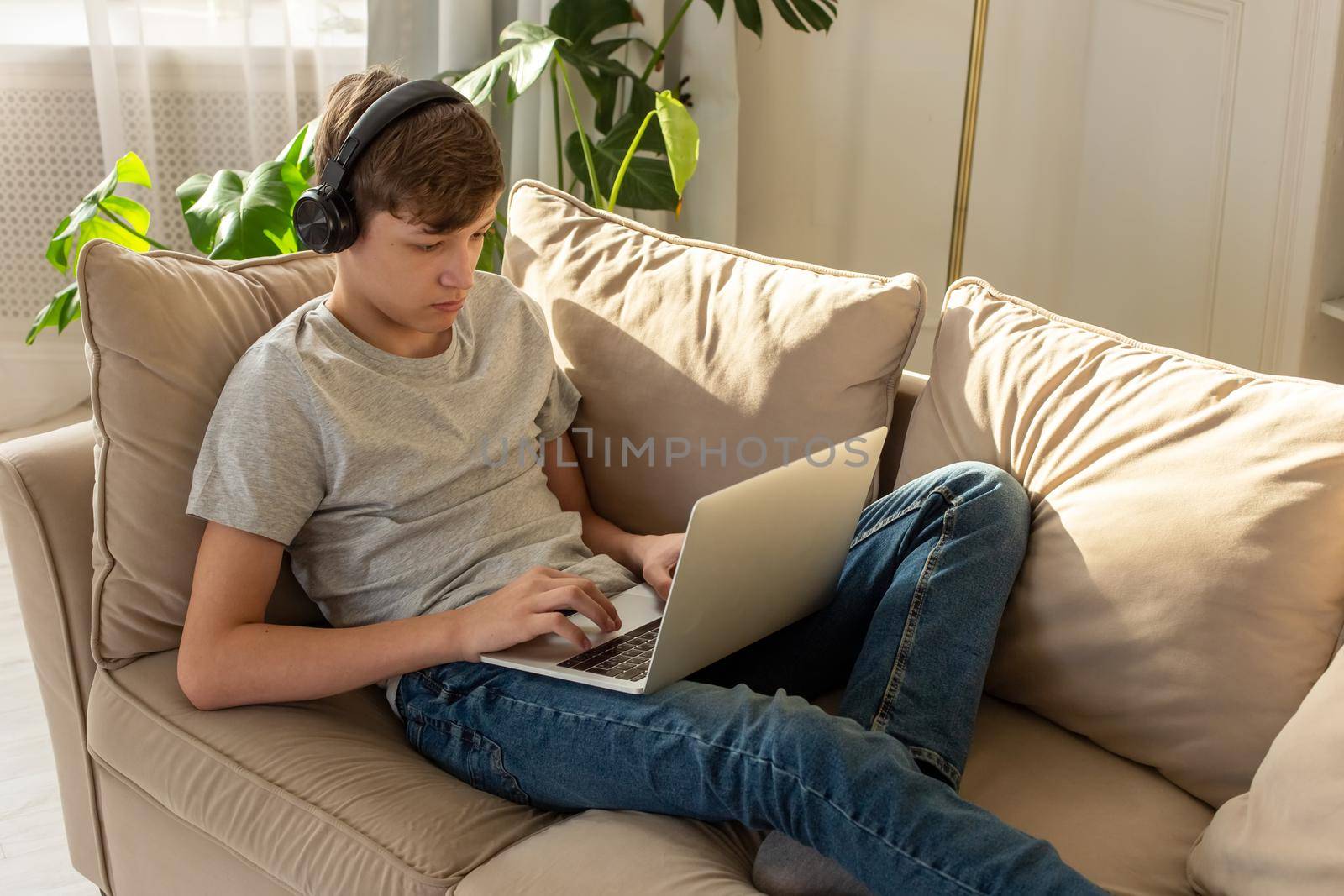 Portrait of teenager boy in a gray t-shirt and blue jeans, lying on a sofa in a room with plant, wearing black headphones on his head, looks into a gray laptop, on his knees. Copy space