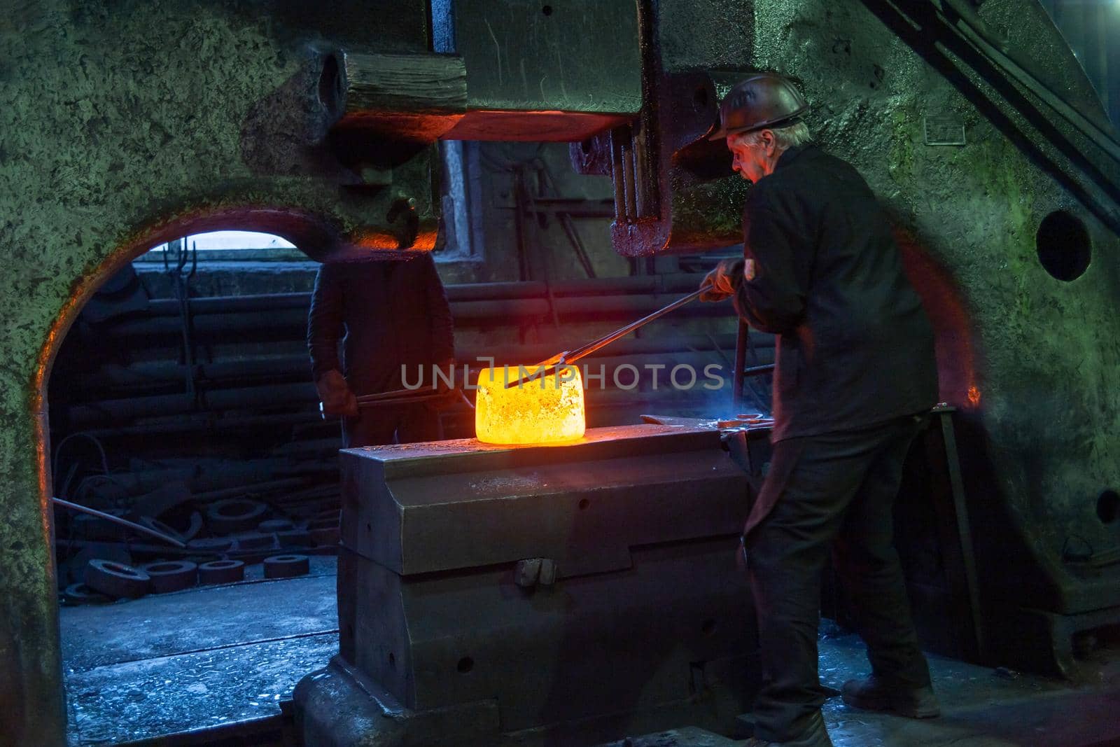 gray haired blacksmith worker taking large piece of hot steel with big forceps in old factory in Tula, Russia - February 1, 2020