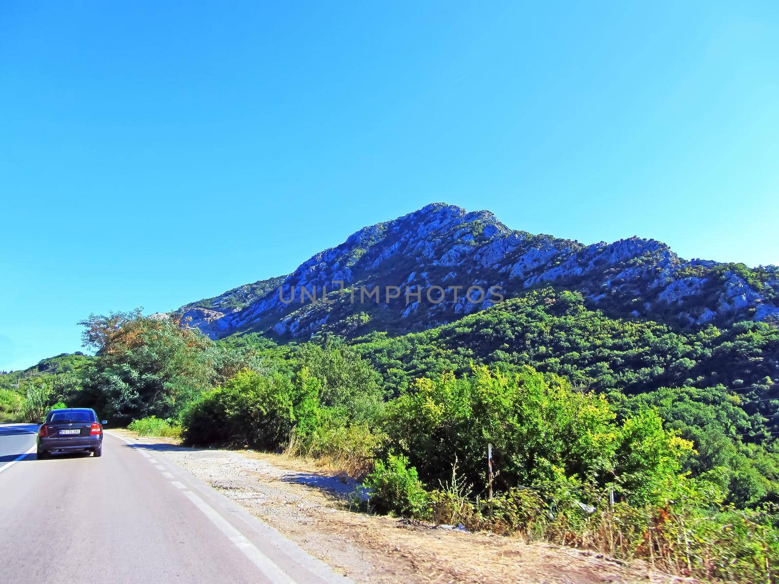 A road with a car and a mountain range in the background. An orga mcala overgrown with greenery in Italy on a summer day. by mr-tigga